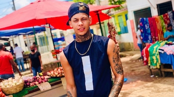 Un joven con gorra oscura y tatuajes posa delante de un puesto de cebollas con carteles de precios bajo sombrillas rojas.