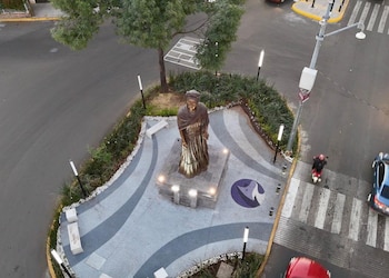 Vista aérea de una escultura de bronce de Frida Kahlo en un pedestal. Está rodeada de jardineras, bancos, farolas y una calle con vehículos