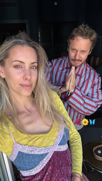 Anita Espasandín en primer plano y Benjamín Vicuña detrás con una chaqueta a rayas y las manos juntas en gesto de saludo