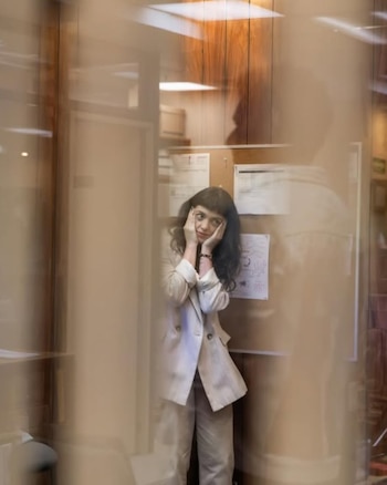 Mujer con cabello oscuro y flequillo, vestida con traje beige, se sujeta la cara con las manos. De pie en una oficina con tablón de anuncios