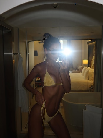 Mujer con traje de baño amarillo de dos piezas y gafas de sol se toma una selfie en un espejo. Se observa un baño, una bañera y un dormitorio reflejado