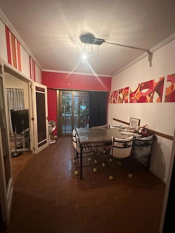 Interior de un comedor con mesa y sillas de hierro forjado, una pared roja y otra con arte abstracto, y una puerta corrediza que da al exterior