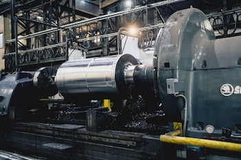 Un torno industrial sostiene una pieza metálica cilíndrica grande junto a acumulaciones de viruta de acero en un taller