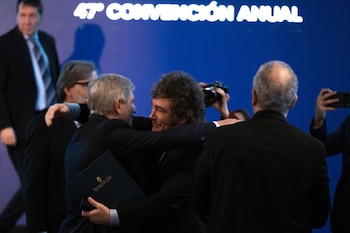 Javier Milei abraza a un hombre con traje frente a una pantalla azul con el texto 47° Convención Anual