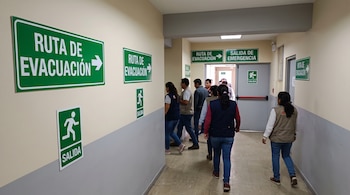 Un grupo de personas camina por un pasillo con carteles verdes de ruta de evacuación y salida de emergencia hacia una puerta gris.