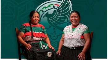 Dos mujeres indígenas sentadas, sonriendo frente a un fondo verde oscuro con un emblema estilizado. Una sostiene una camiseta de fútbol negra de México.
