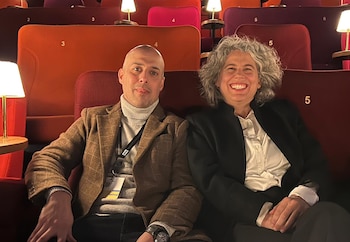 Un hombre calvo con chaqueta a cuadros y una mujer de pelo gris rizado sonriente, sentados en butacas rojas de cine con pequeñas lámparas laterales