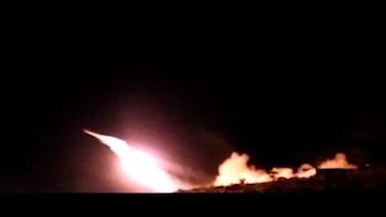 Un resplandor brillante traza una diagonal hacia el cielo nocturno con humo y fuego en el suelo