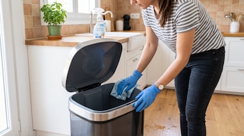 Una persona con guantes azules pasa un paño por el interior de un cubo de basura metálico abierto junto a una encimera de cocina.