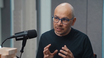 Primer plano de un hombre calvo con gafas azules y barba corta, vestido con una camiseta negra, gesticulando con sus manos frente a un micrófono Shure.