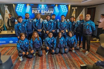 Un grupo de trece atletas y entrenadores con chándales azul y negro posa frente a una pantalla con el logo de la Copa Internacional de Lucha Olímpica Pat Shaw