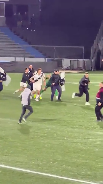 Futbolistas con camiseta blanca y personas de particular corren por la cancha de fútbol junto a agentes de seguridad con escudos