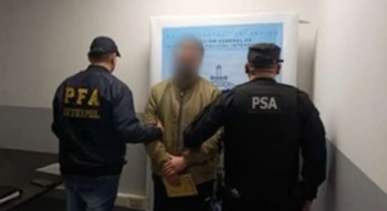 Un hombre con el rostro difuminado está de pie entre dos agentes de policía vestidos de azul y negro frente a un cartel