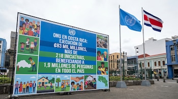 Gran cartel azul y verde detalla iniciativas de la ONU en Costa Rica, con ilustraciones y texto, junto a las banderas de la ONU y Costa Rica en un entorno urbano.