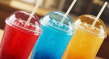 Primer plano de tres vasos transparentes con tapas de domo y pajitas, conteniendo bebidas heladas de colores rojo, azul y amarillo con hielo.