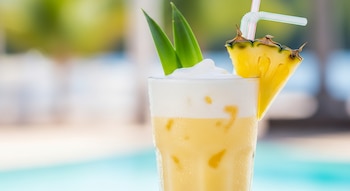 Primer plano de un vaso alto de piña colada con hielo, una capa de espuma blanca, una rodaja de piña, dos hojas verdes y una pajita rayada.