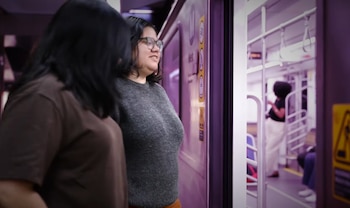 Dos mujeres de pie junto a la puerta abierta de un tren; una lleva gafas y un suéter gris, sonríe hacia el interior, mientras otra persona está dentro
