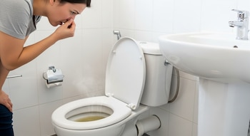 Mujer inclinada sobre un inodoro con orina amarillenta y vapor, cubriéndose la nariz. En un baño blanco, se ve también un lavamanos y un rollo de papel.