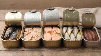 Cinco latas de conservas de pescado abiertas, que muestran caballa, atún, salmón, sardinas y anchoas, sobre una mesa de madera.