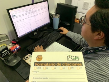 Un hombre con gafas y camisa de rayas trabaja en un escritorio; se ve una mano en el teclado y otra sosteniendo un formulario de citaciones con logo PGN