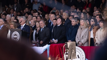 Javier Milei en un podio con micrófonos, junto a una multitud de personas. Se observa el logo de la Sociedad Rural Argentina y una bandera argentina