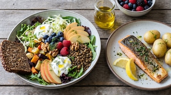 Primer plano de una comida que incluye un plato de salmón con limón y patatas, y un tazón grande de ensalada con bayas, nueces y pan.