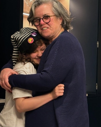 Una mujer adulta con cabello gris y lentes abraza a una persona joven con un gorro de rayas blanco y negro y tres insignias de colores