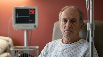 Hombre mayor en bata de hospital, sentado en habitación roja. Monitor médico con trazado cardíaco y glóbulos rojos conceptuales. Soporte de goteo visible.