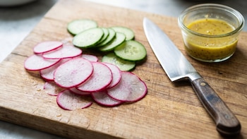Rodajas de rábano y pepino, un cuchillo con mango de madera y un cuenco de vidrio con aderezo sobre una tabla de cortar de madera