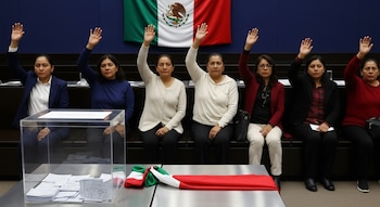 Seis mujeres sentadas levantan la mano frente a una bandera de México; una urna transparente con boletas y una banda tricolor se observan en primer plano.