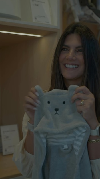 Una mujer sonriente, de cabello largo, sostiene una prenda de bebé gris con capucha de oso. Lleva un anillo, un reloj y una pulsera. Fondo difuso con estanterías