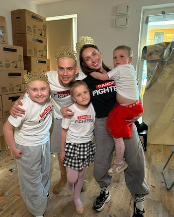 Una familia de cinco (dos adultos, tres niños, incluyendo un bebé) sonríe con camisetas 'Cancer Fighters' y coronas doradas, entre cajas de mudanza y confeti