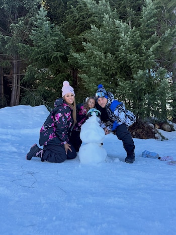 Un hombre, una mujer y una niña posan rodeando un muñeco de nieve sobre la superficie nevada de un bosque de pinos