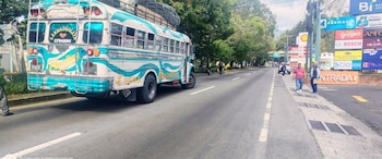 Autobús blanco y celeste con nombres pintados y carga en el techo, personas, carretera, árboles, postes, múltiples señales comerciales y un letrero de entrada