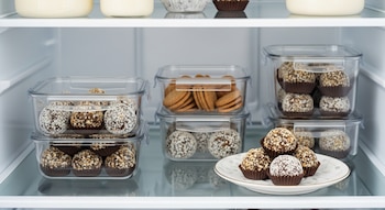 Trufas de galletitas y leche condensada dentro de recipientes transparentes apilados en una nevera, con algunas trufas sobre un plato blanco al frente.