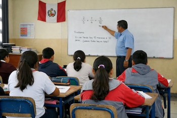 Un profesor de pie junto a una pizarra con problemas de matemáticas, cuatro alumnos sentados de espaldas a la cámara en pupitres de madera, una bandera de Perú.