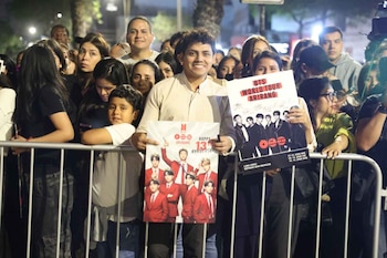 Un hombre joven sonríe mientras muestra dos carteles de BTS detrás de una barandilla metálica rodeado por una multitud de personas