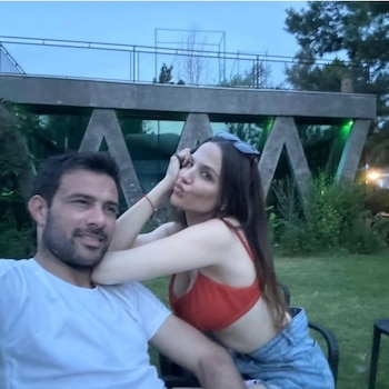 Un hombre y una mujer posan juntos al aire libre. Ella, con top rojo, se apoya en su hombro haciendo un puchero; él sonríe. Hay vegetación de fondo