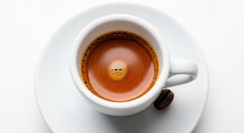Vista cenital de una taza de espresso blanca con café oscuro y crema rojiza. Un grano de café descansa en el platillo blanco sobre un fondo blanco.