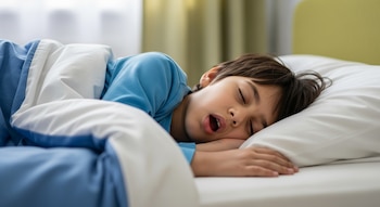 Niño duerme de costado en cama con sábanas blancas y manta azul. Tiene boca abierta y cabello oscuro. Cortinas y pared clara de fondo.