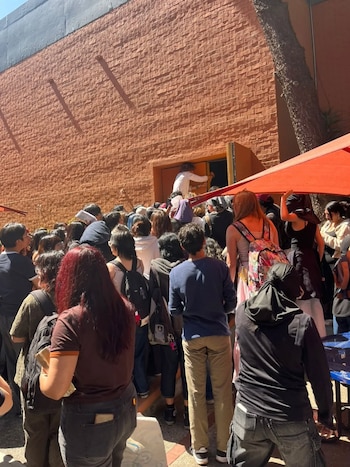 Gran multitud de estudiantes se congrega frente a la entrada de un edificio de ladrillo bajo un toldo rojo, con un árbol visible a la derecha