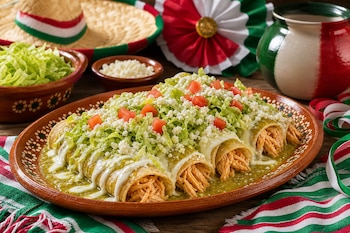 Un plato de barro servido con enchiladas verdes cubiertas de crema, lechuga, queso y tomate, rodeado de adornos mexicanos en una mesa de madera.