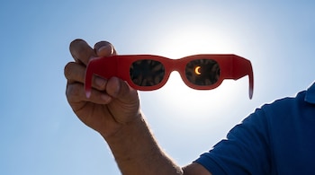 Unas gafas rojas de eclipse solar son sostenidas en alto contra un cielo azul claro, revelando una forma de luna creciente del sol en sus lentes oscuras.