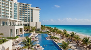 Vista aérea de un gran hotel blanco con múltiples piscinas frente a una playa de arena blanca, palmeras y el mar Caribe turquesa bajo un cielo azul claro.