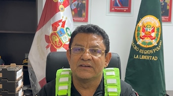Un hombre con anteojos y chaleco reflectante verde se encuentra ante banderas de la Policía Nacional y de Perú en una oficina