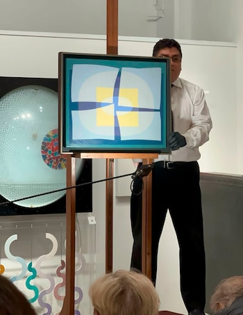 Hombre con camisa blanca, corbata y guantes negros presenta una obra de arte abstracta enmarcada sobre un caballete ante una audiencia
