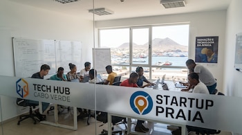 Oficina moderna con jóvenes adultos trabajando en laptops. Pizarras blancas, ventana grande con vista a un puerto, barcos y montañas. Letreros de 'Tech Hub Cabo Verde' y 'Startup Praia'.