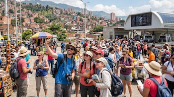 Una multitud de turistas extranjeros con sombreros y cámaras toma fotos frente a una estación de MetroCable y casas de ladrillo en una colina de Medellín.