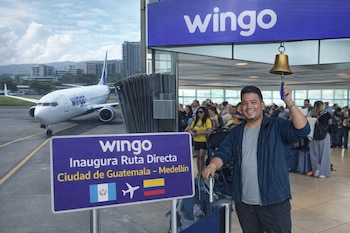 Un hombre sostiene una campana dorada junto a un cartel de Wingo con banderas de Guatemala y Colombia, un avión y personas en un aeropuerto.