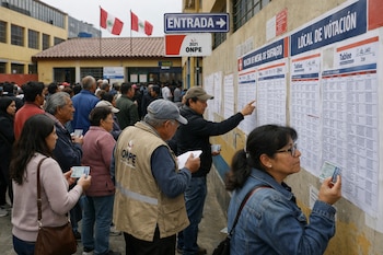 Una multitud de personas de diversas edades en fila frente a una pared con carteles de mesas de votación. Muchos sostienen su DNI. Se ven banderas peruanas y señalización ONPE.